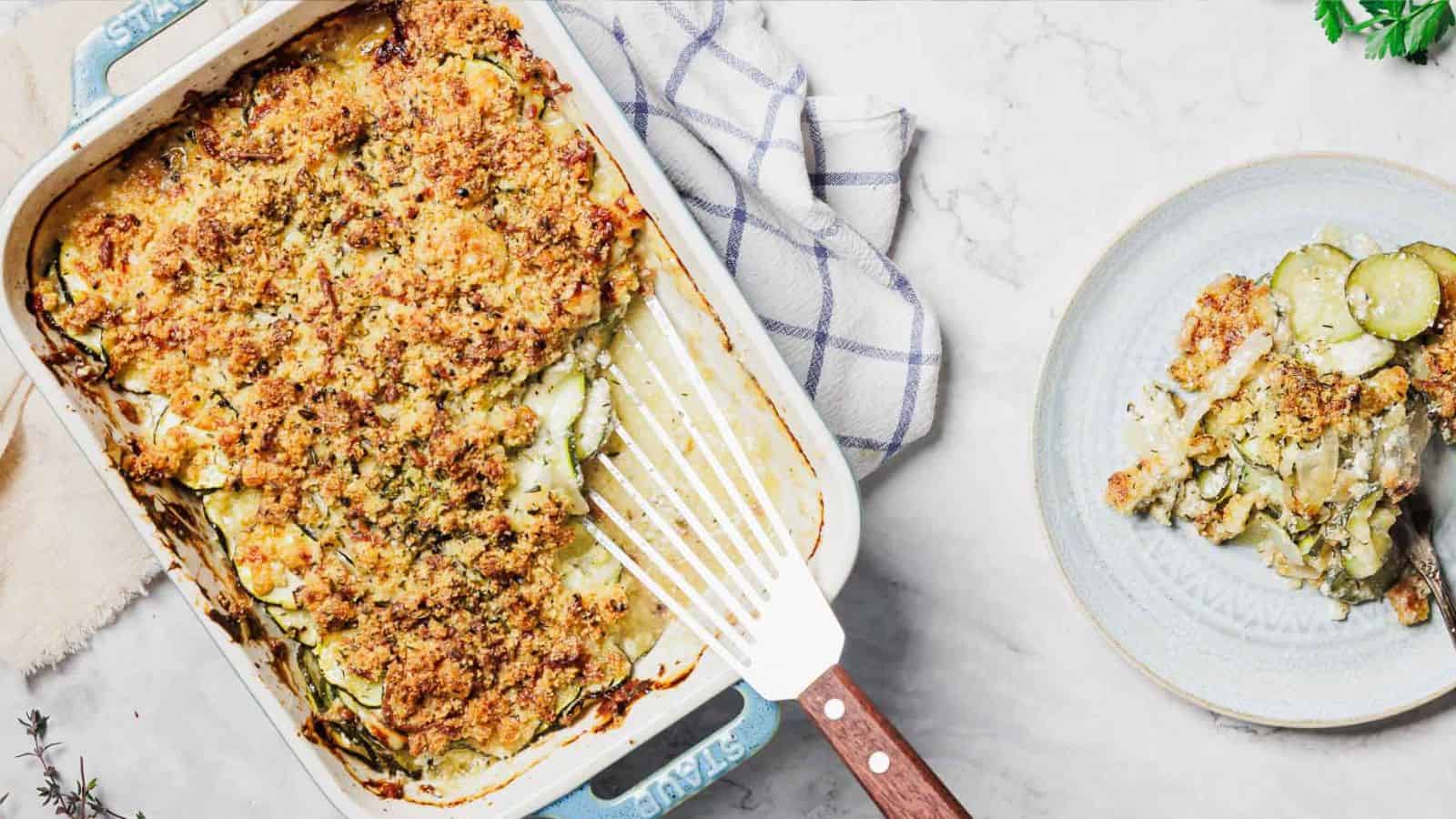 A baked dish with a golden-brown crumb topping is in a rectangular baking dish, which has a white and blue checkered cloth nearby. A serving is on a round plate to the right. The surface is a light marble texture.