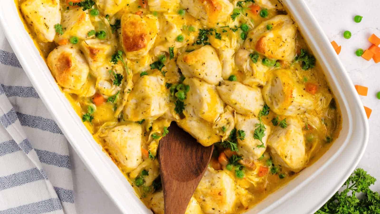 A casserole dish filled with baked chicken, mixed vegetables, and creamy sauce, with a wooden spoon resting inside.