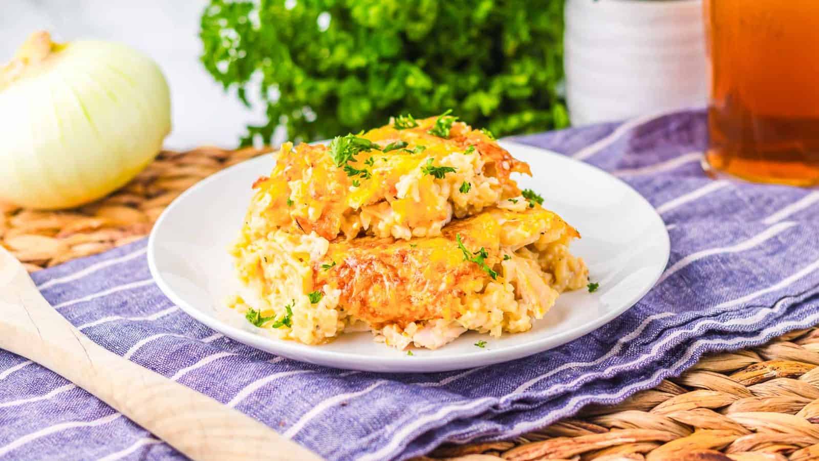 A white plate holds a serving of cheesy casserole garnished with chopped parsley, placed on a blue-striped cloth next to a wooden spoon, an onion, and green leafy parsley in the background.