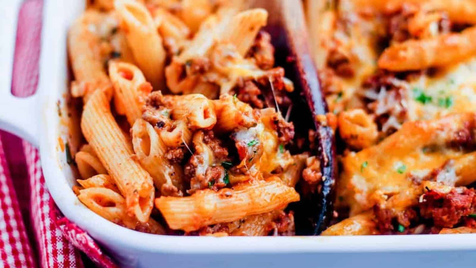 A wooden spoon scooping a portion of penne pasta bake.