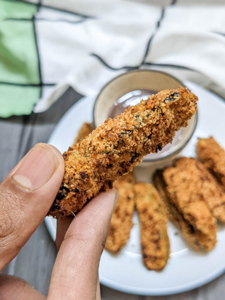 One Air fried pickle slice held in a hand in focus with other slices in the background blurred on a white plate with a bowl of dip by the side with a green and white cloth napkin visible in the background.