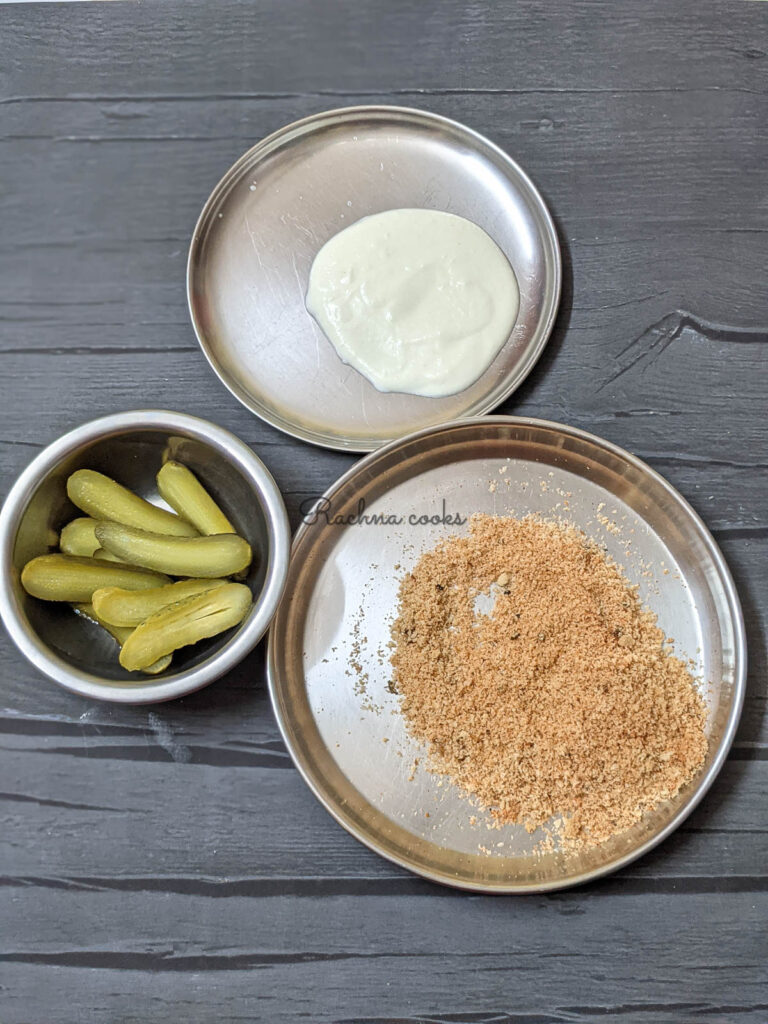 3 shallow plates visible. One plate has breadcrumb mixture, 2nd one has yogurt and flour slurry and third one has pickles sliced in half.